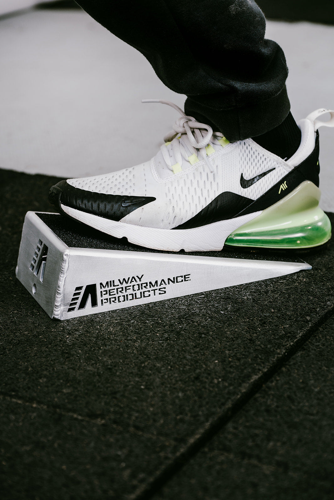 Akan Milway wearing white sneakers using the slant board to stretch ankles and calves with 'Milway Performance Products' branding visible.